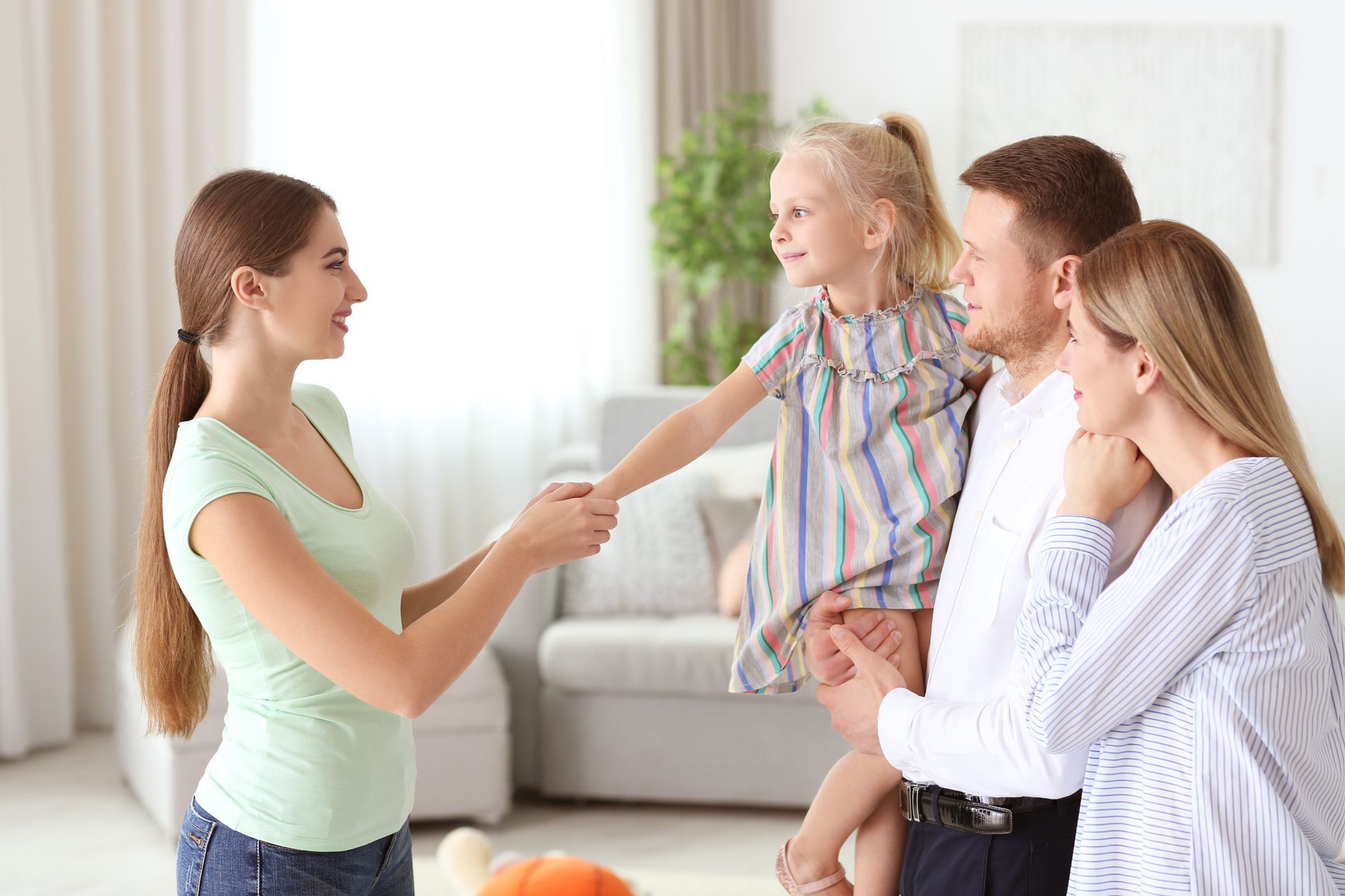 Woman holds hands with a young girl as the girl's parents watch, smiling. Indoor, bright room.