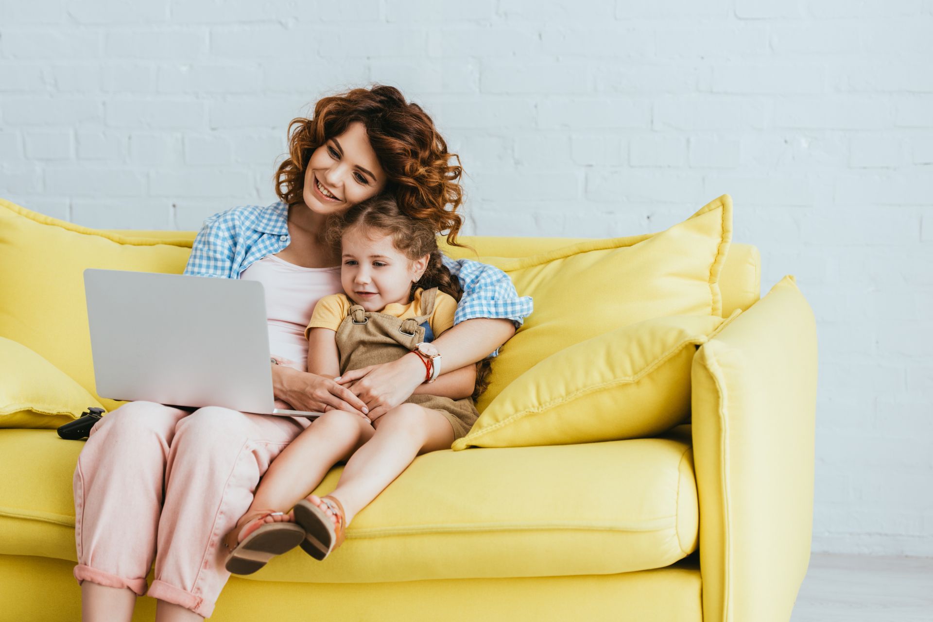 Woman and girl sit on a yellow couch, looking at a laptop. Both are smiling.