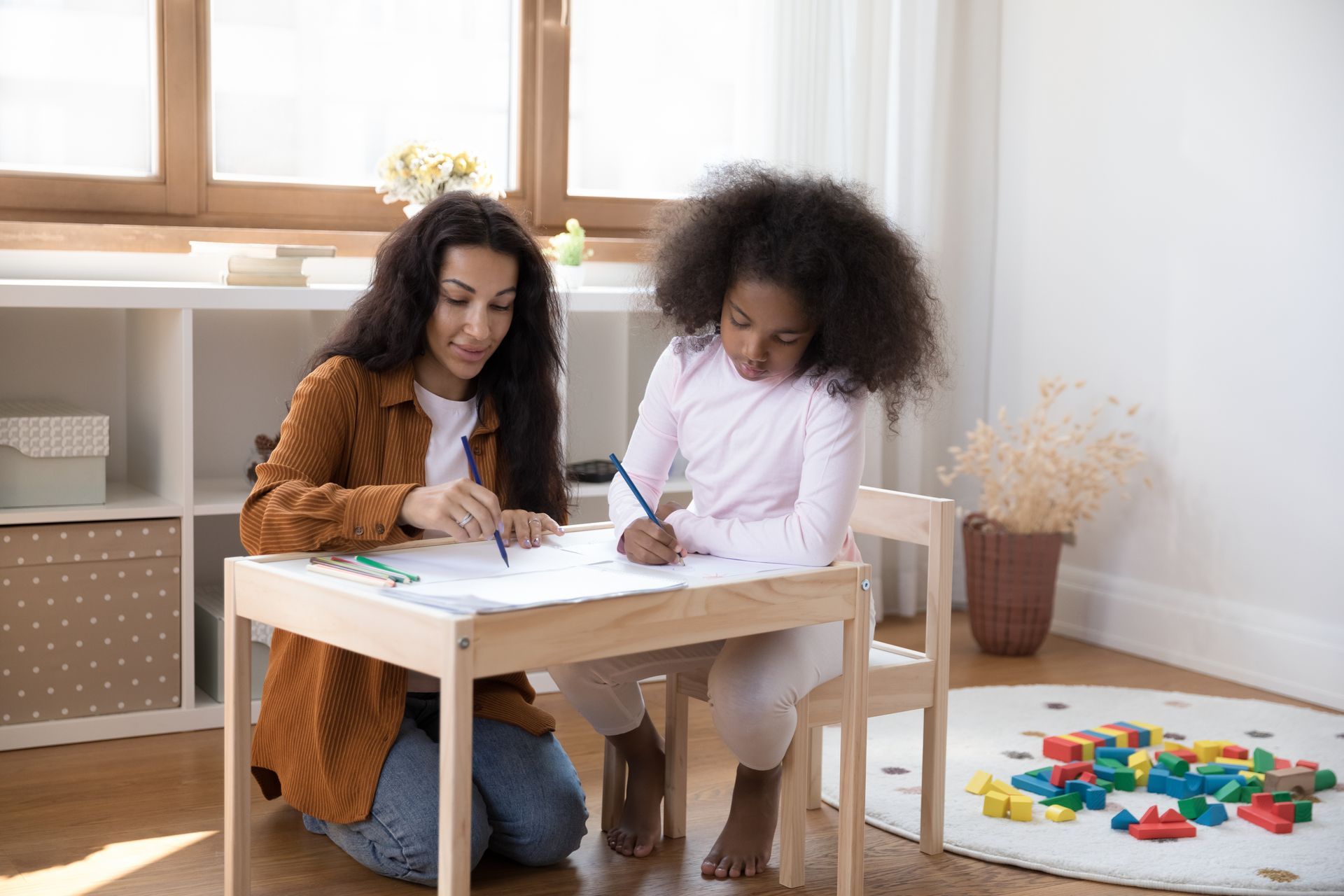 Woman and child drawing at a table indoors. The woman kneels, while the child sits, both smiling.