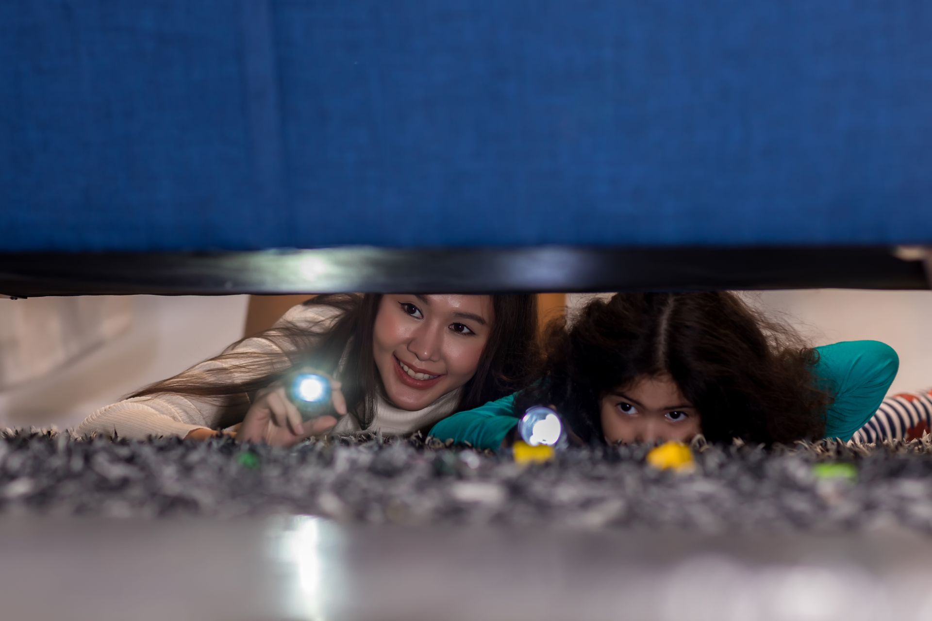 Woman and child with flashlights peering under a bed, smiling and looking for something.