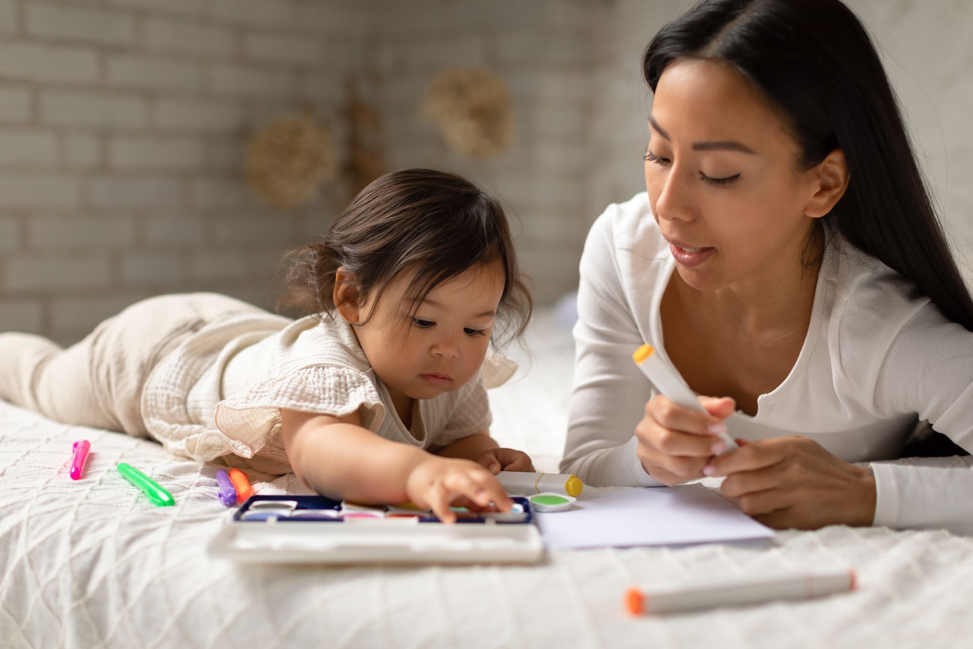 Woman and child coloring on a bed. Woman holds a marker, child points at crayons.