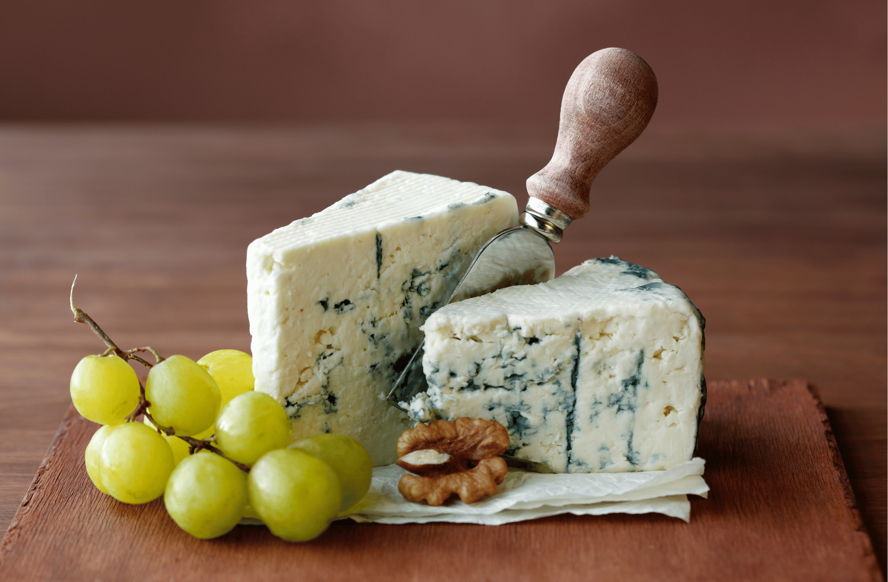 A piece of blue cheese is sitting on a wooden cutting board next to grapes and walnuts.