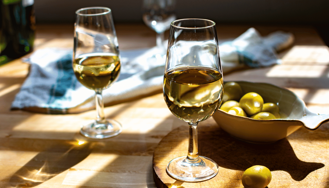 Two glasses of white wine are sitting on a wooden table next to a bowl of olives.