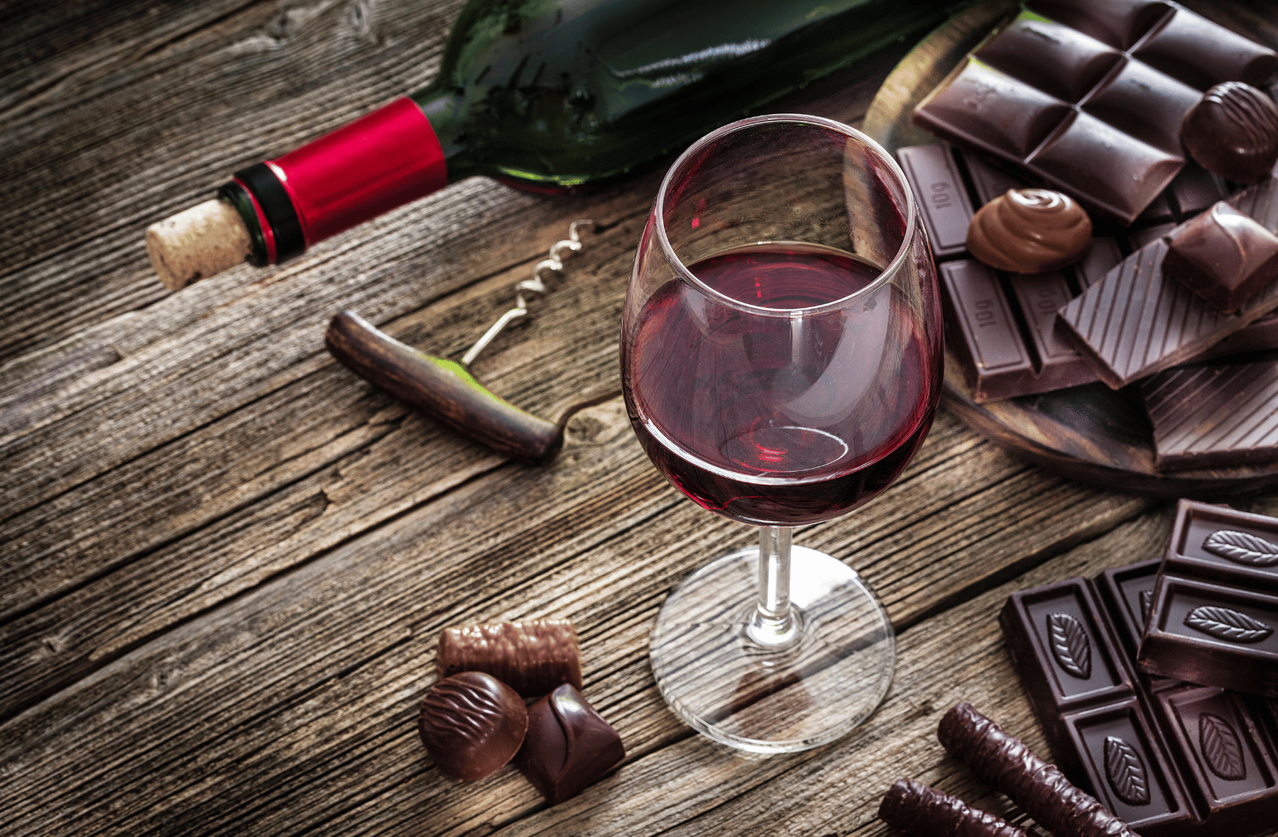 A bottle of wine , a glass of wine , chocolate bars and a corkscrew on a wooden table.