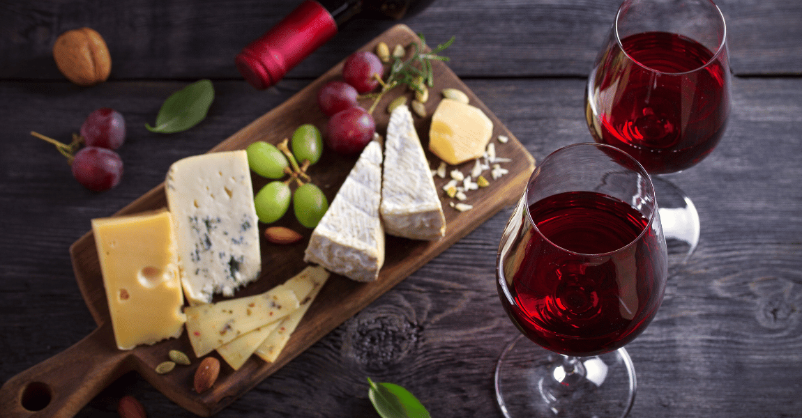 A wooden cutting board topped with cheese , grapes , nuts and two glasses of red wine.