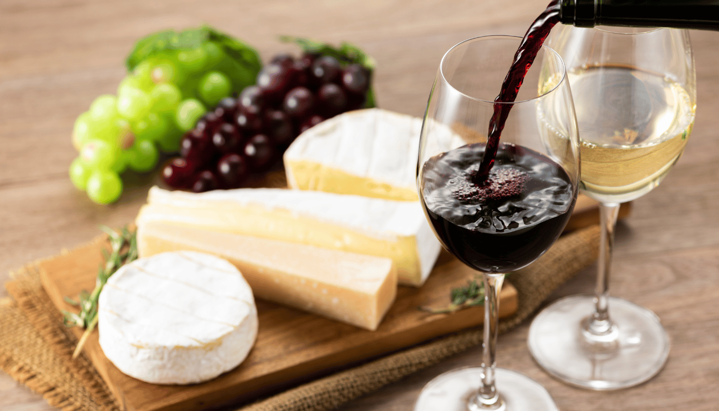 A bottle of wine is being poured into a glass next to a cutting board with cheese and grapes.