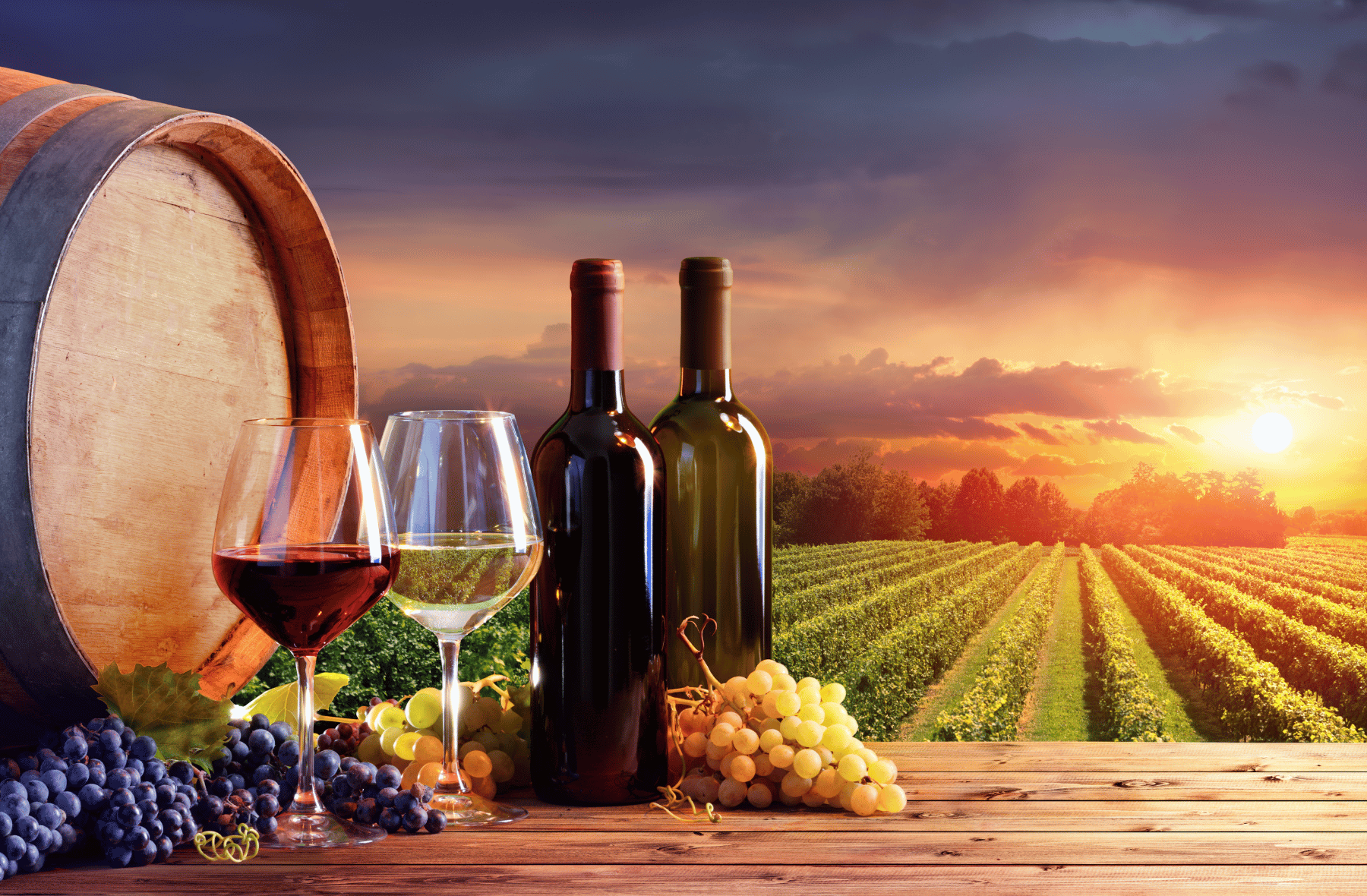 A wooden table topped with bottles of wine , glasses of wine , grapes and a barrel.