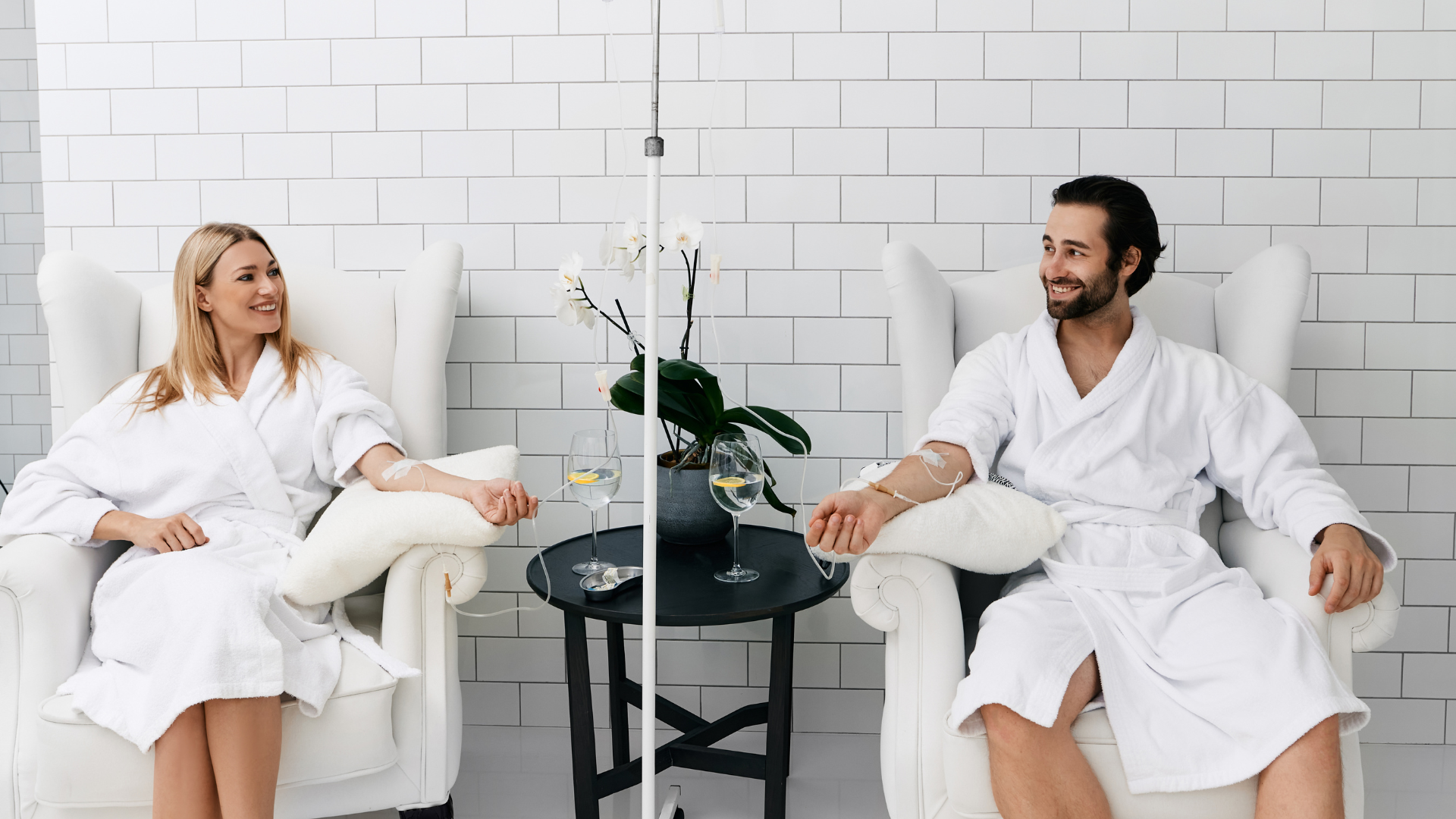 Two people in white robes sit in chairs, receiving IV drips. They smile at each other, near a small table.