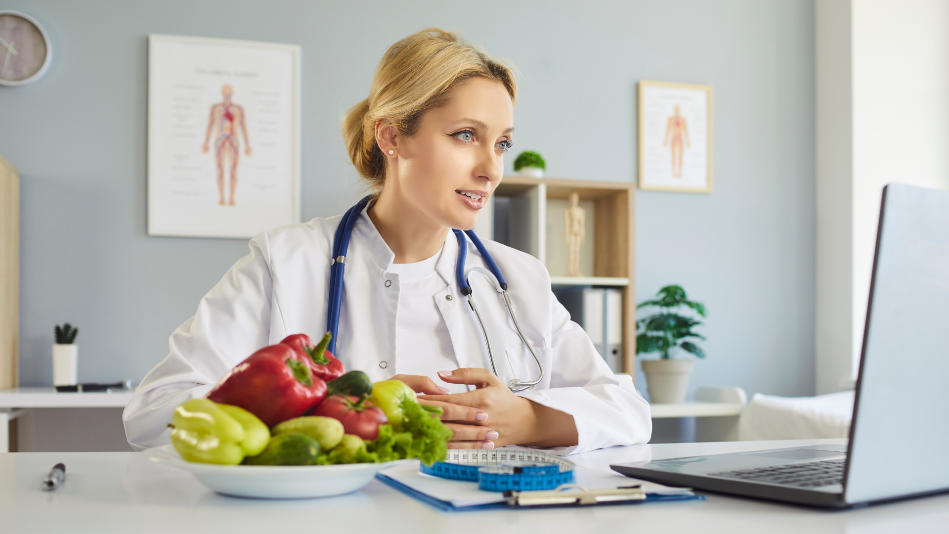 A nutritionist wearing a lab coat and stethoscope speaks via video call behind a desk with a bowl of fresh vegetables.