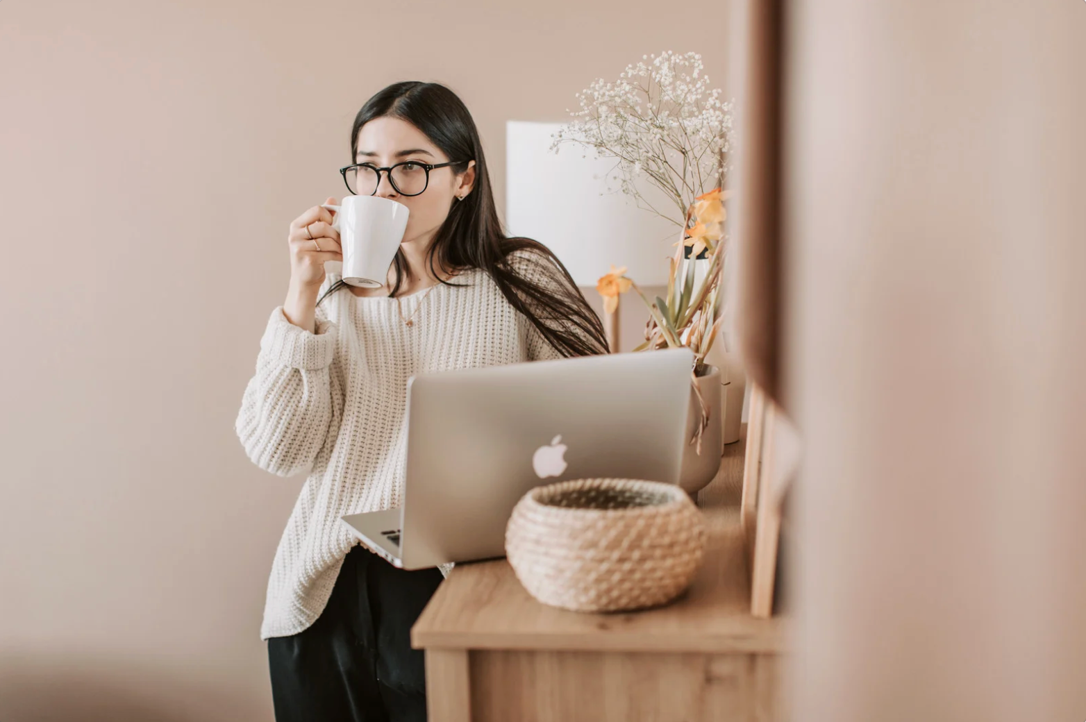 Woman drinking from a mug while leaning against a wooden table with a laptop, neutral background.