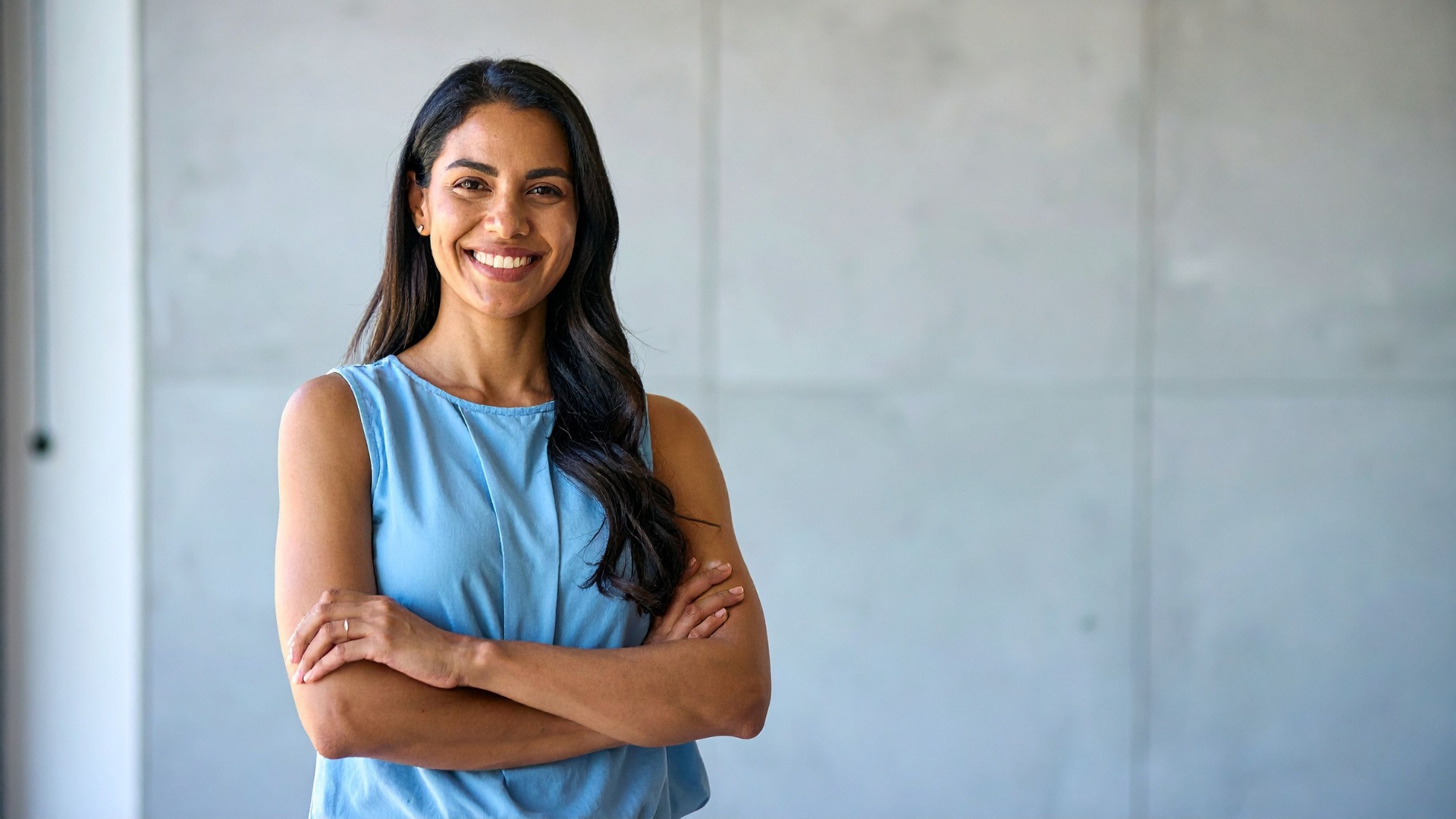 A smiling person in a blue sleeveless top stands with their arms crossed against a plain grey concrete background.