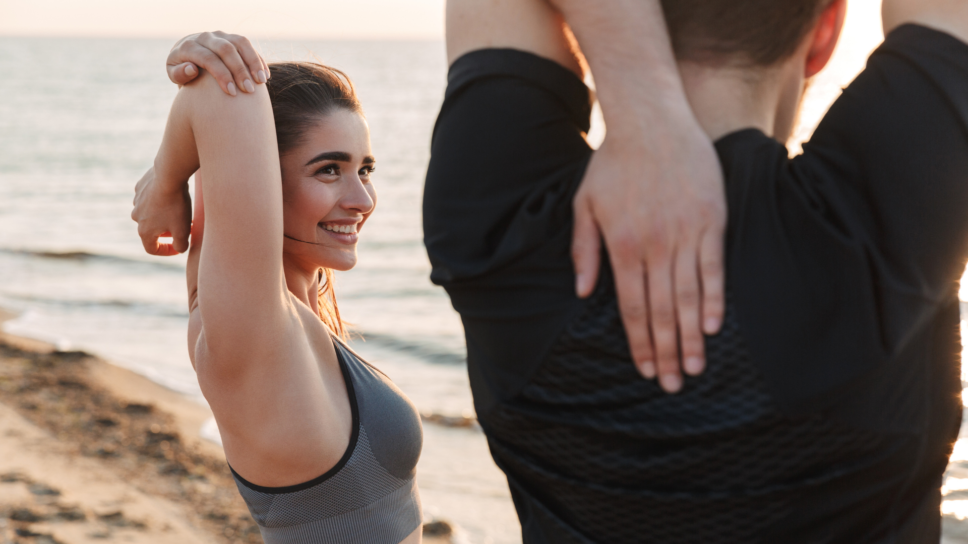 Two people stretching their arms above their heads while standing on a beach at sunset.