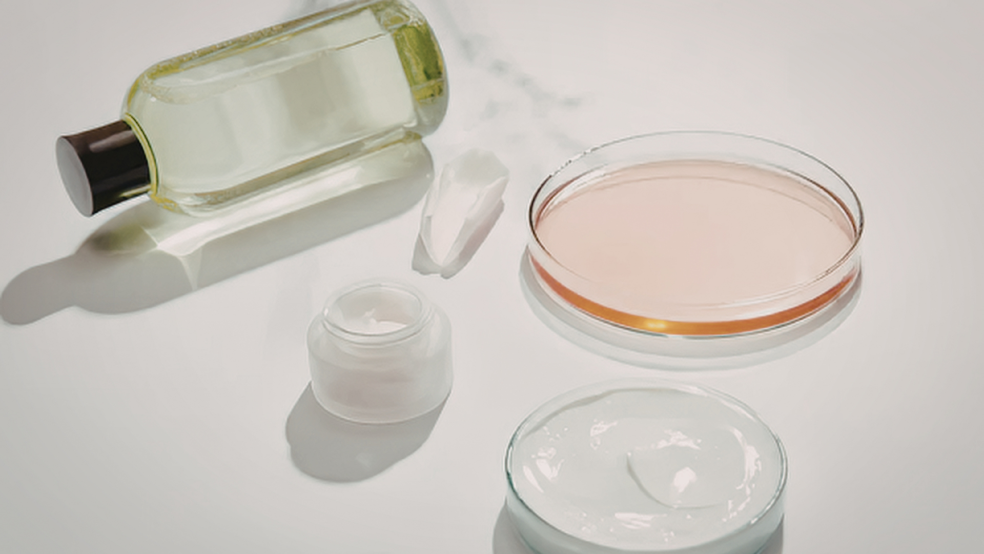 Skincare products: a clear bottle of liquid, a small cream jar, and two petri dishes containing serum and white cream.