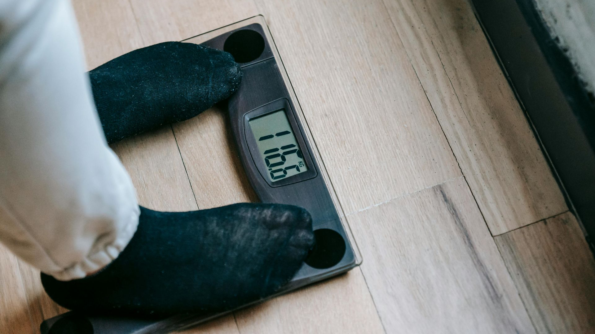 Person's feet on a digital scale, showing a weight of 116.1 pounds. Wearing black socks, on a wood floor.
