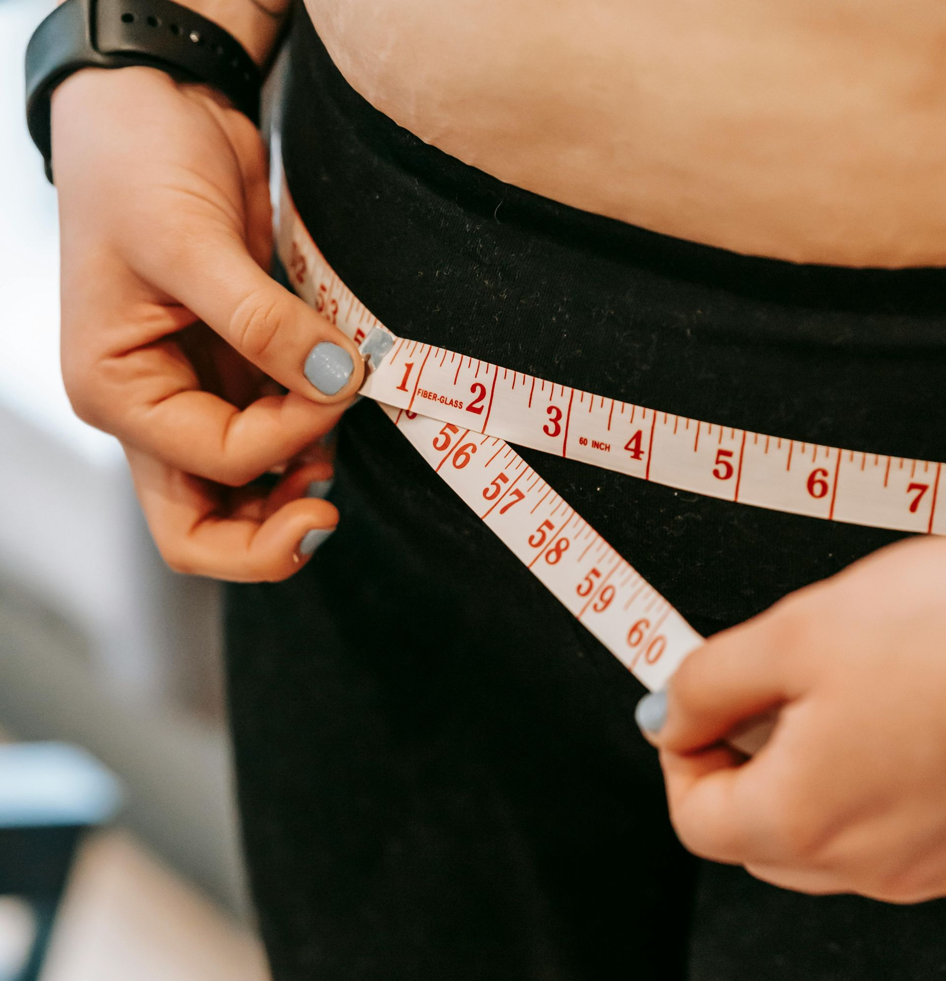 Person measuring waist with a white measuring tape, wearing black pants, black watch, light blue nail polish.