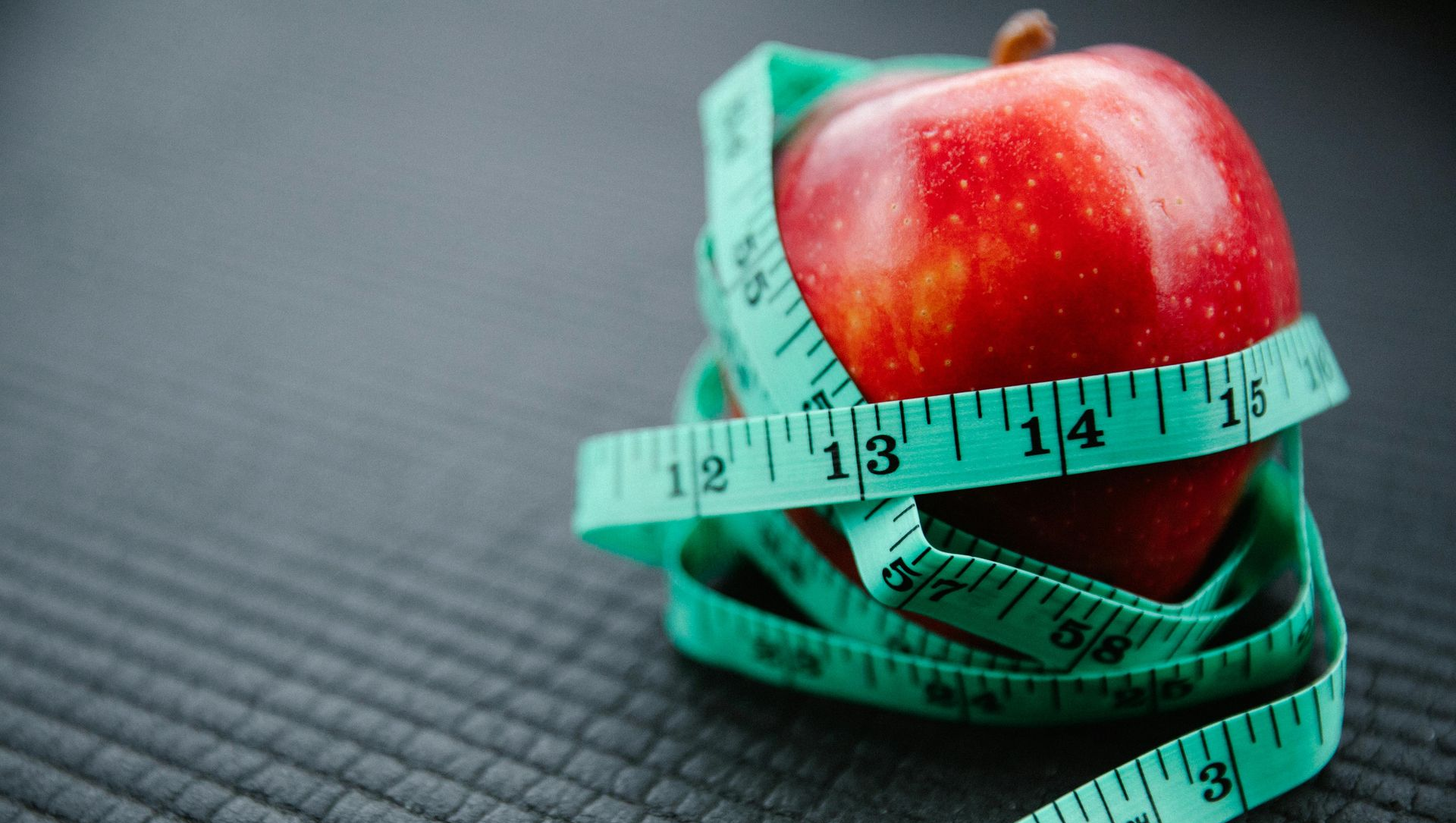 Red apple wrapped in a green measuring tape, on a gray surface.