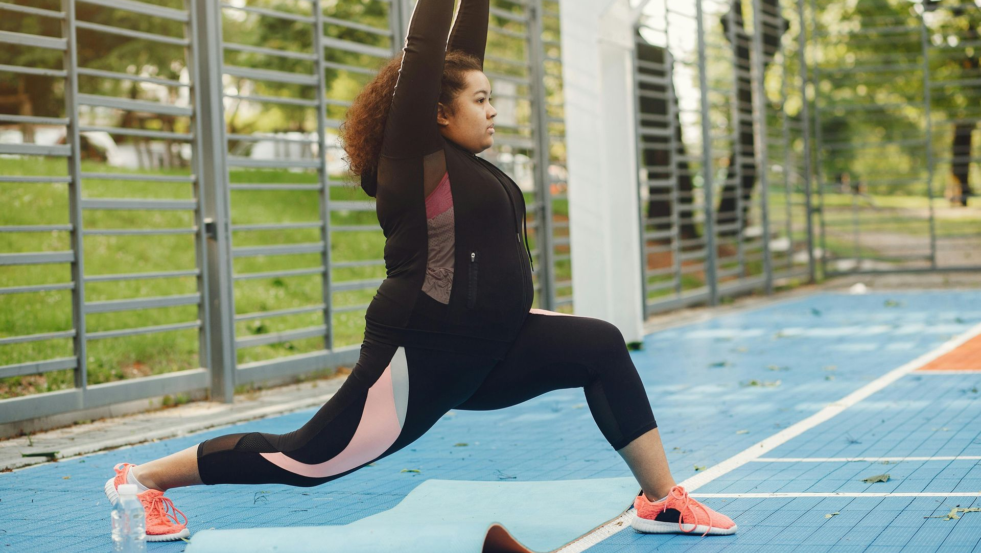 Woman in workout gear lunging on blue mat outdoors, arms raised.