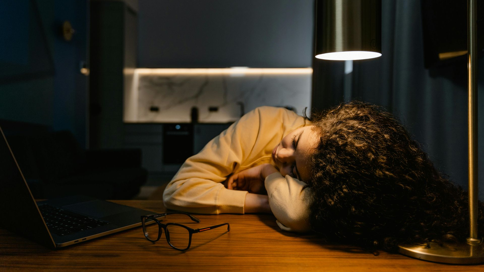 Woman asleep at a desk in dim room, head on arms, near a laptop and glasses.