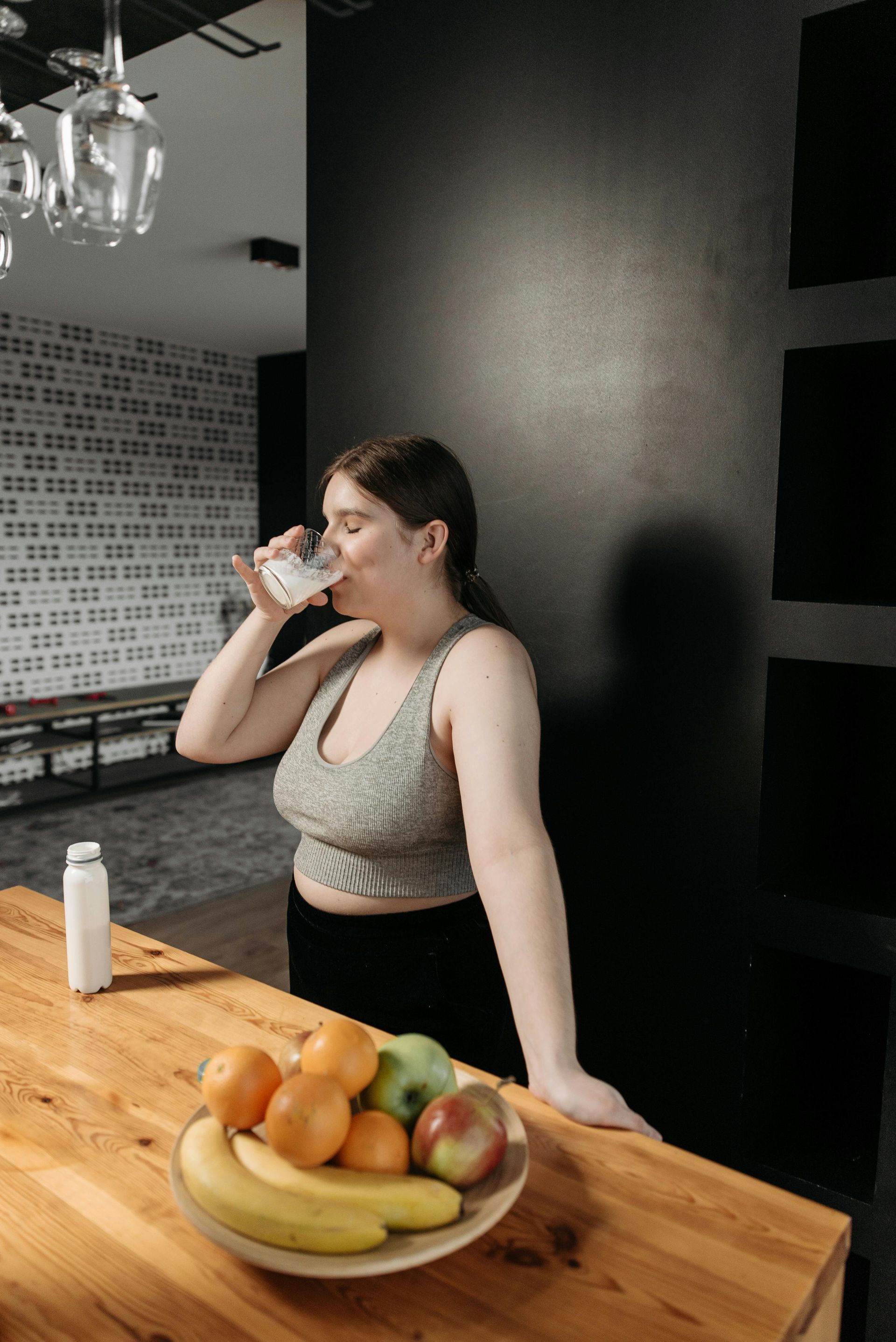 Woman drinking water by a fruit bowl and black wall.
