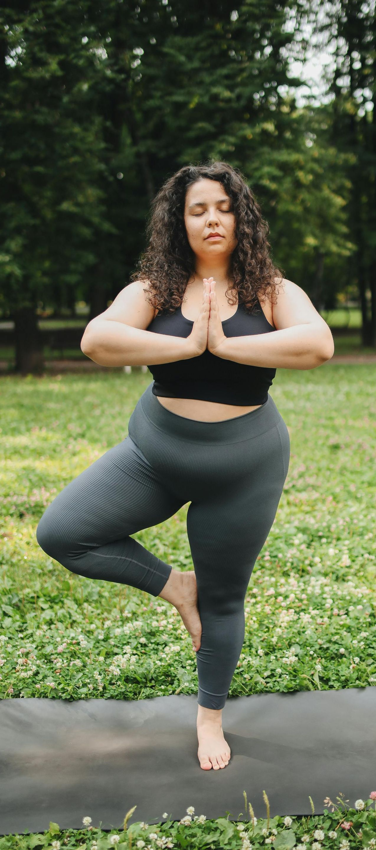 Woman doing yoga tree pose outdoors, green grass, closed eyes, dark leggings, black top.