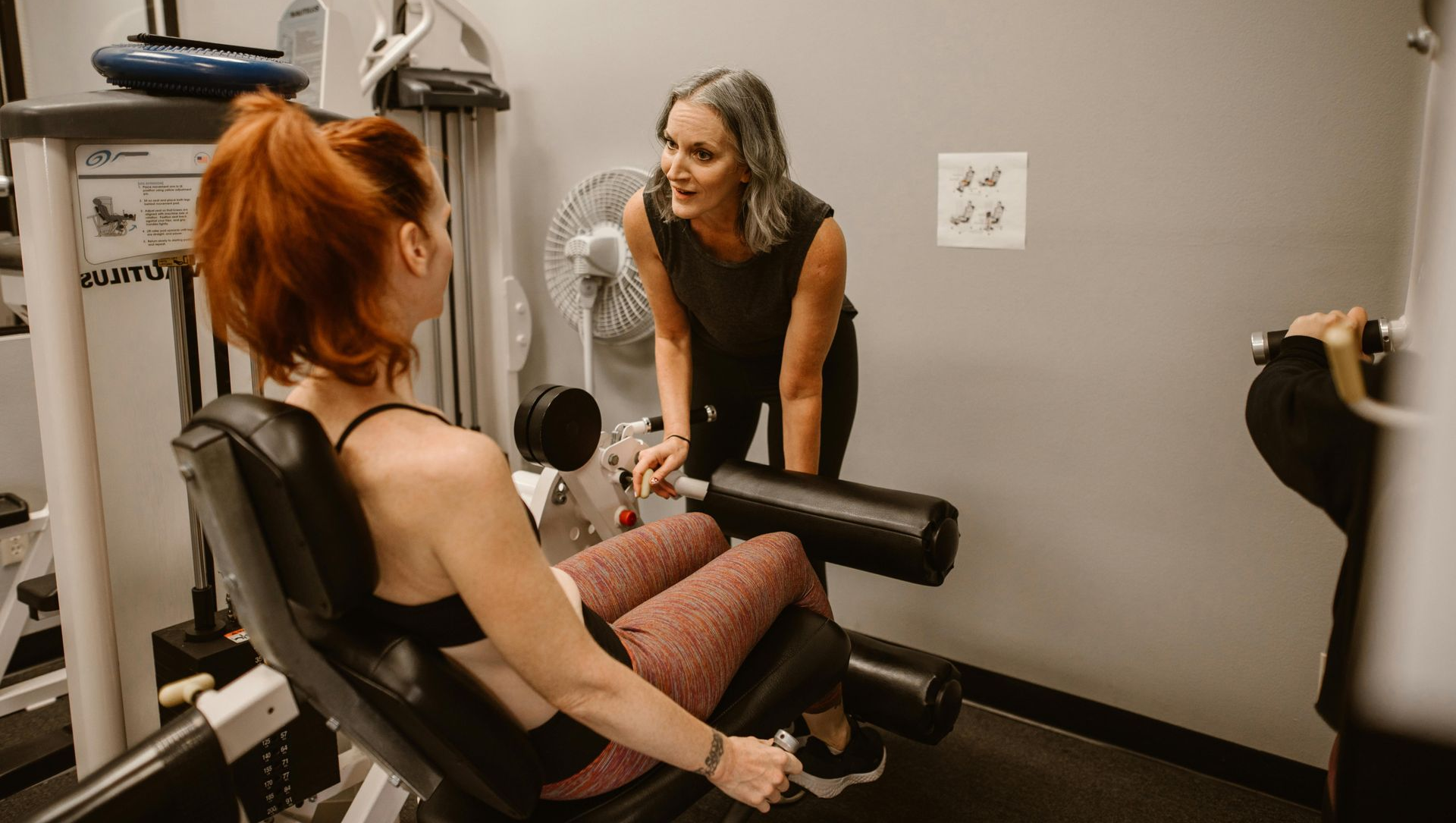 Woman exercising legs on a machine, being coached by a woman in a gym.