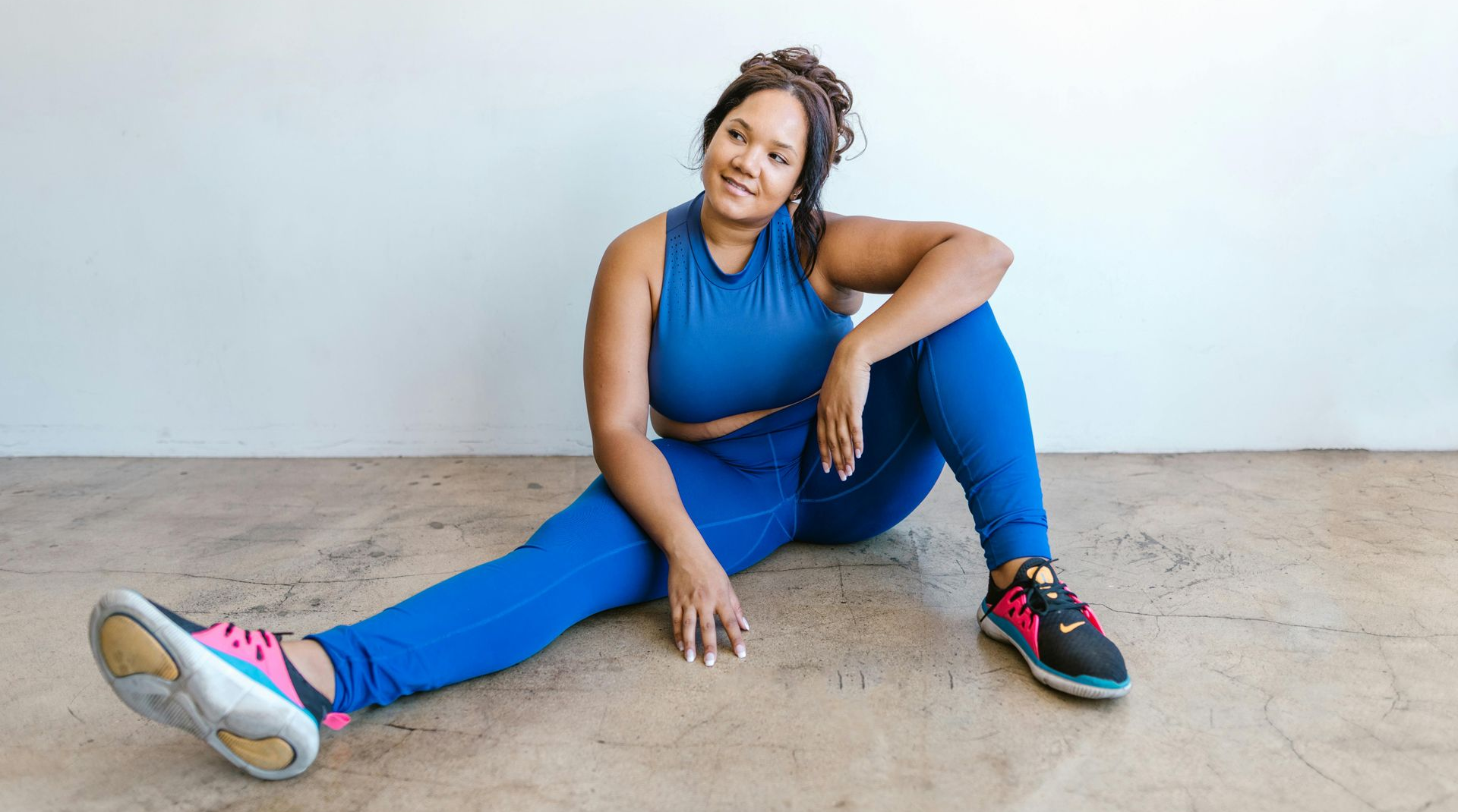 Woman in blue workout clothes sits on the floor, smiling. Leaning on arm, legs extended.