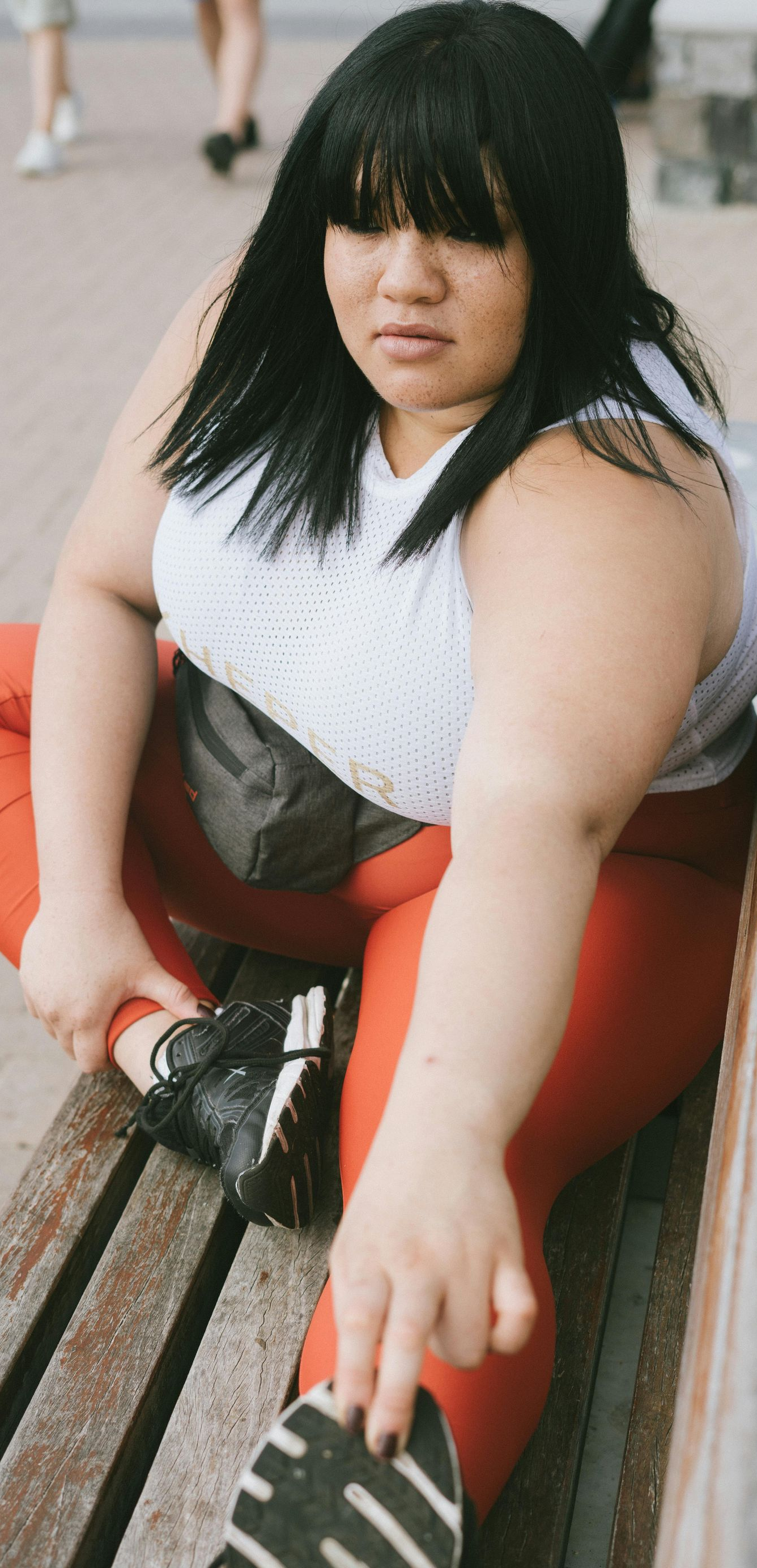 Woman with black hair in white top and red pants, stretching on a bench outdoors.