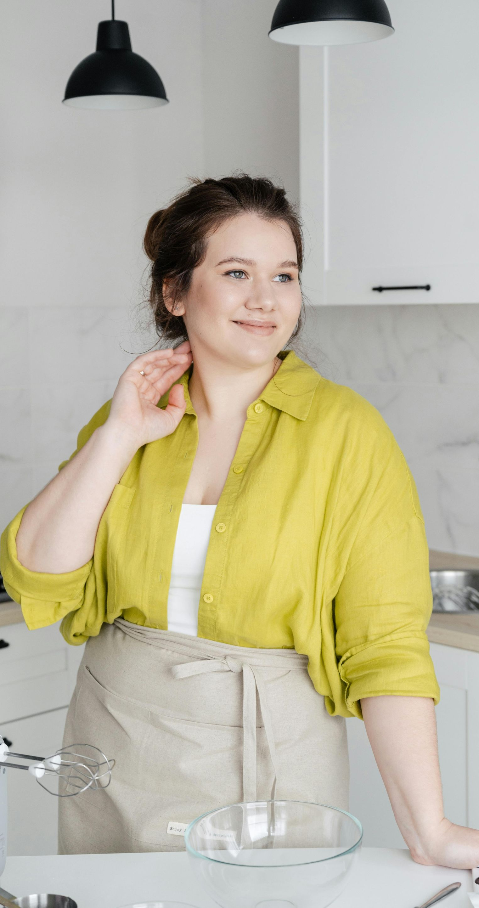 Woman in kitchen, wearing an apron, smiling, hand near neck.  Bright yellow shirt, white top.