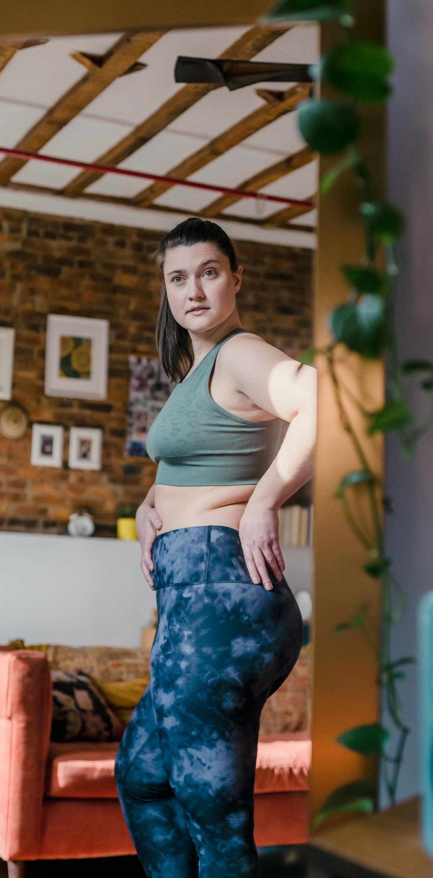 Woman in workout clothes leans against a mirror, posing in front of a brick wall interior.