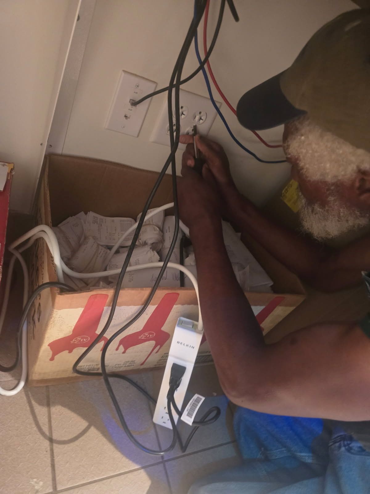 Man in cap wiring electrical outlet. Wires and power strip visible.