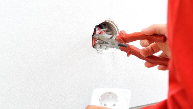 Person in red sleeves using pliers on an electrical outlet in white wall.