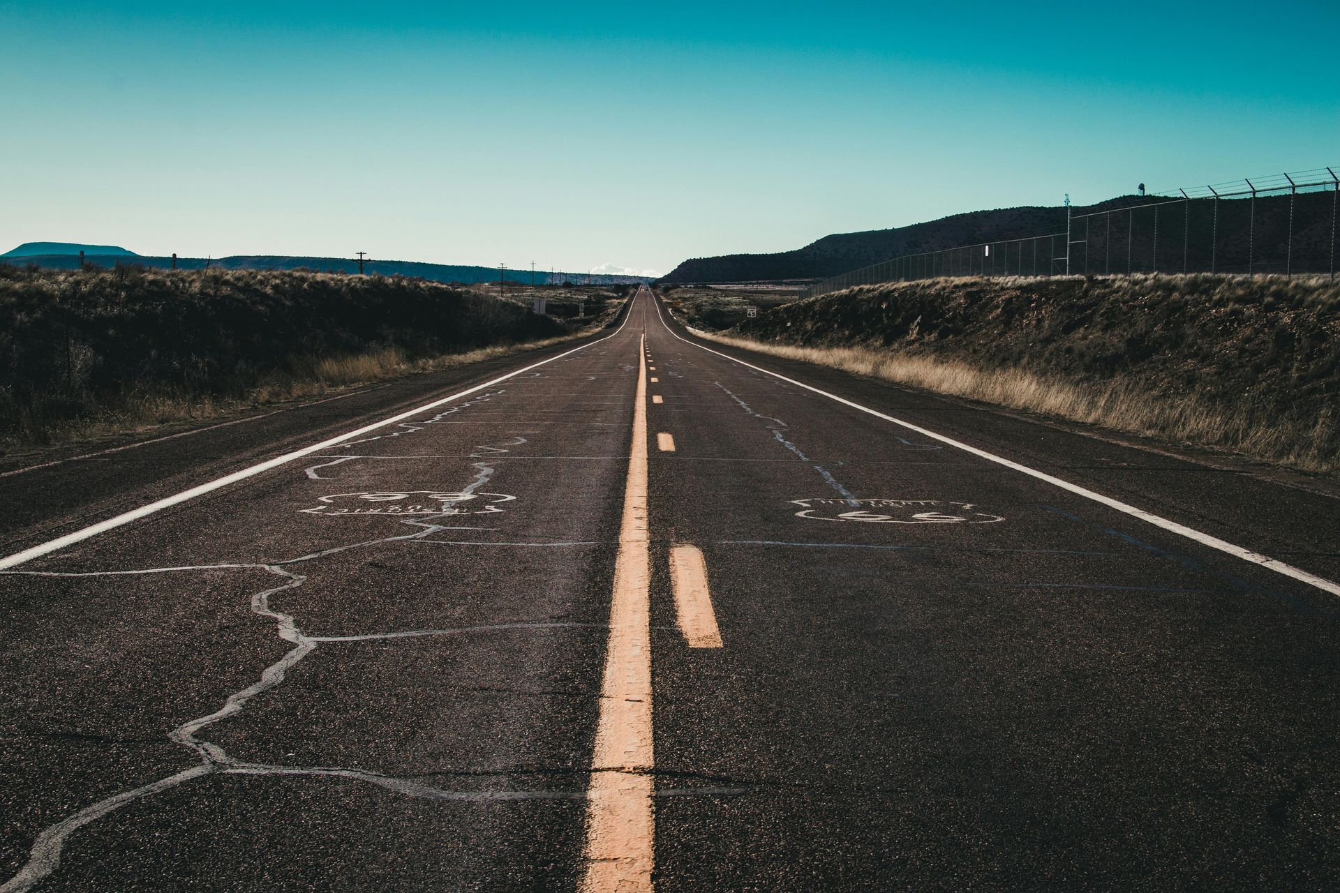 Empty two-lane road stretching into the distance under a clear sky, with fields on both sides.