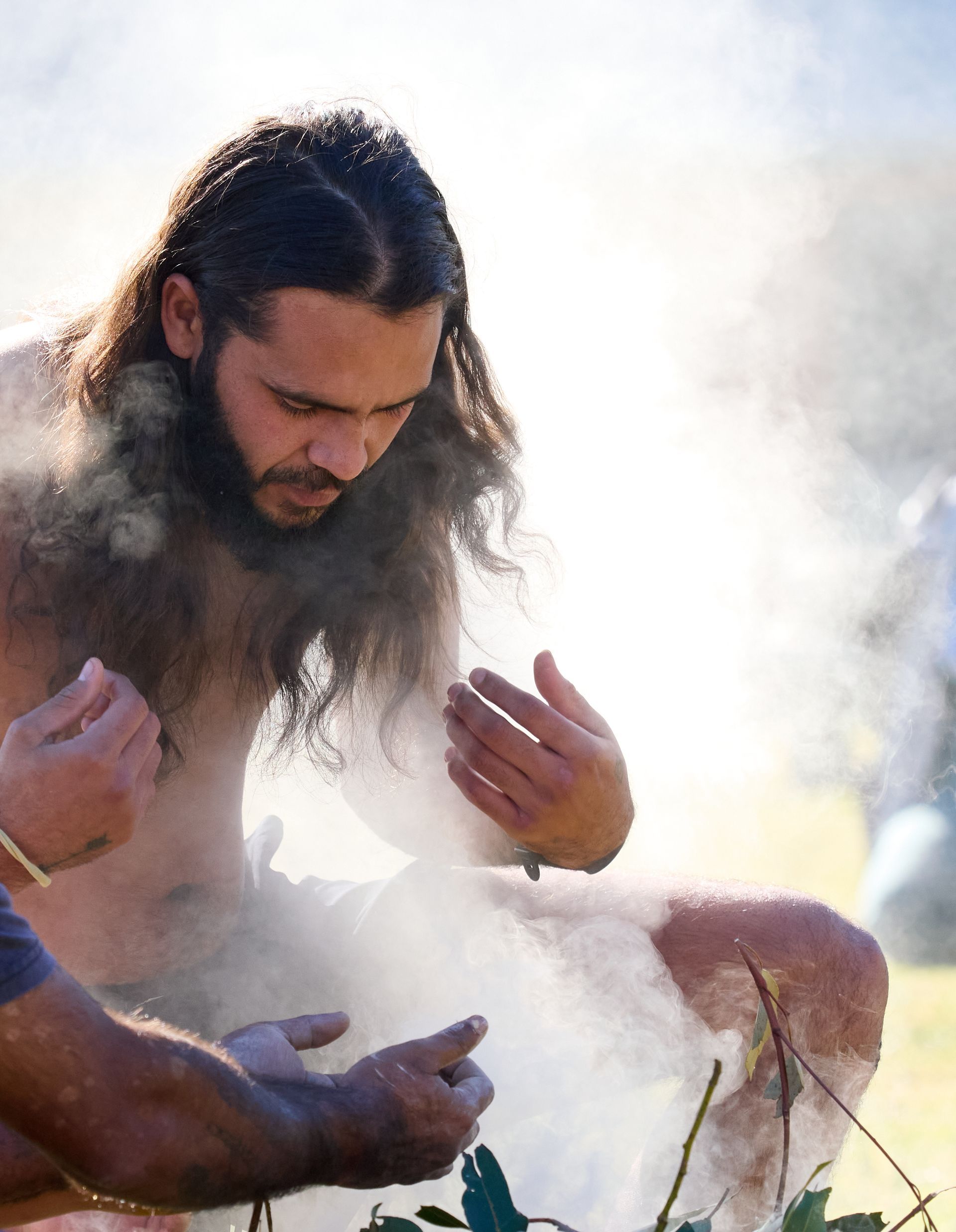 Man with long hair and beard, shirtless, seated in smoke, looking down. Outdoors.