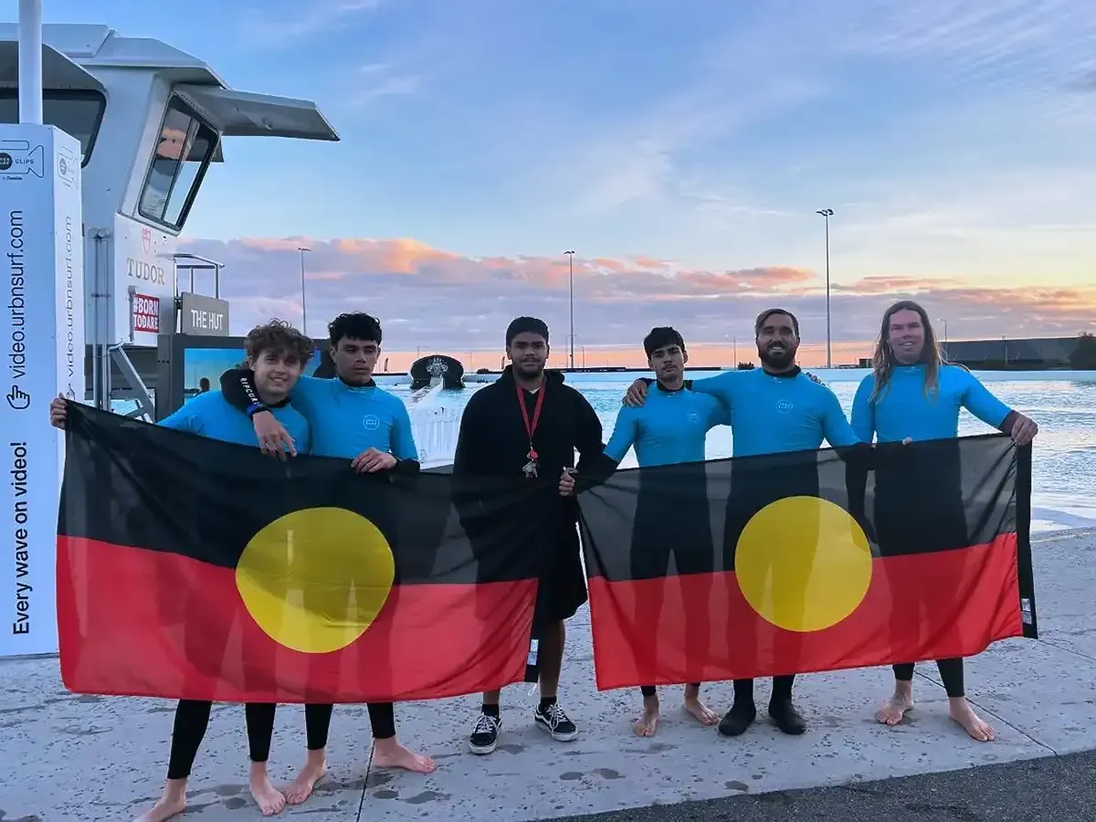 A group holds an Aboriginal flag at a water park, smiling, wearing blue tops.