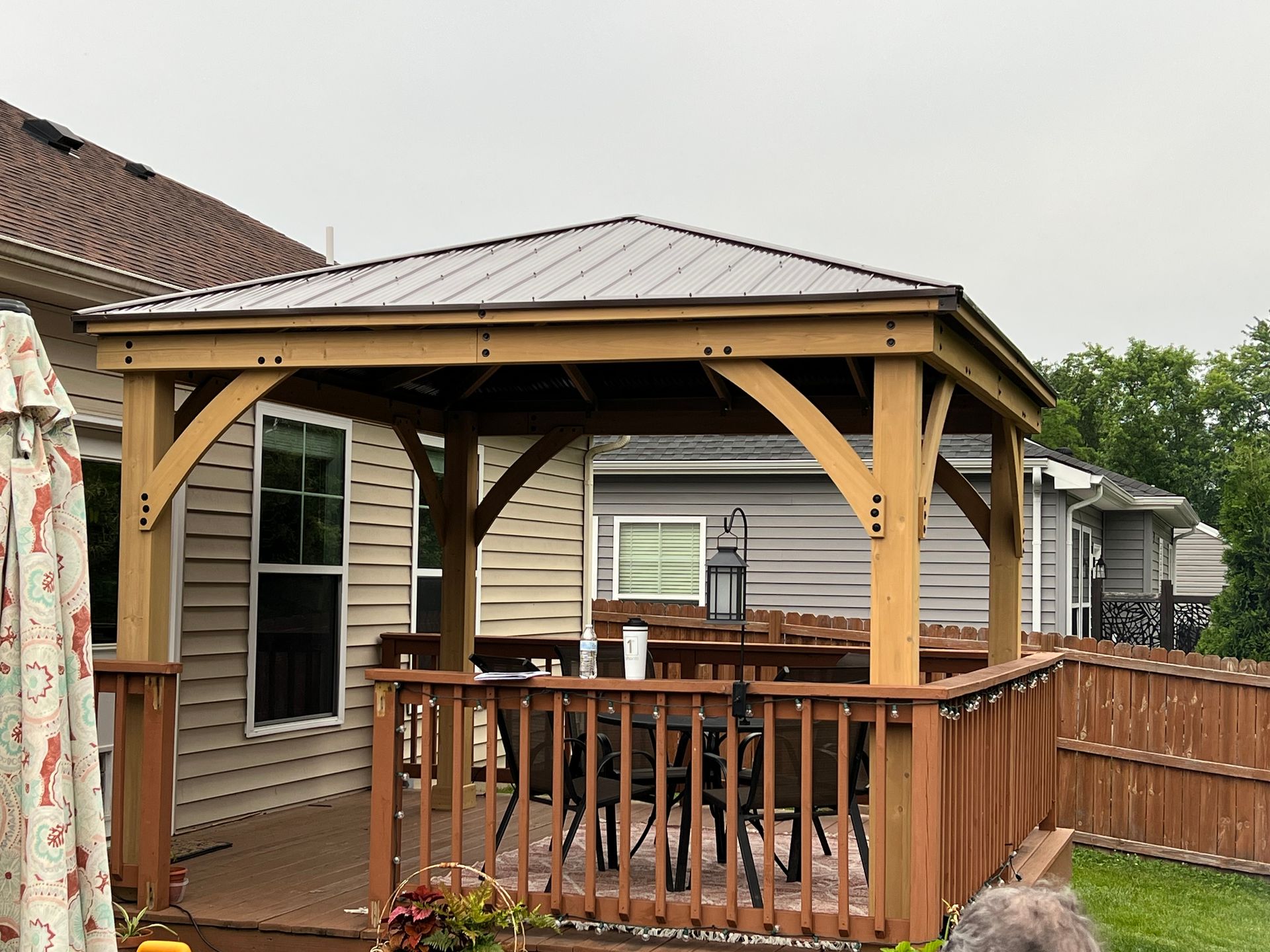 A wooden gazebo with a metal roof is in the backyard of a house.