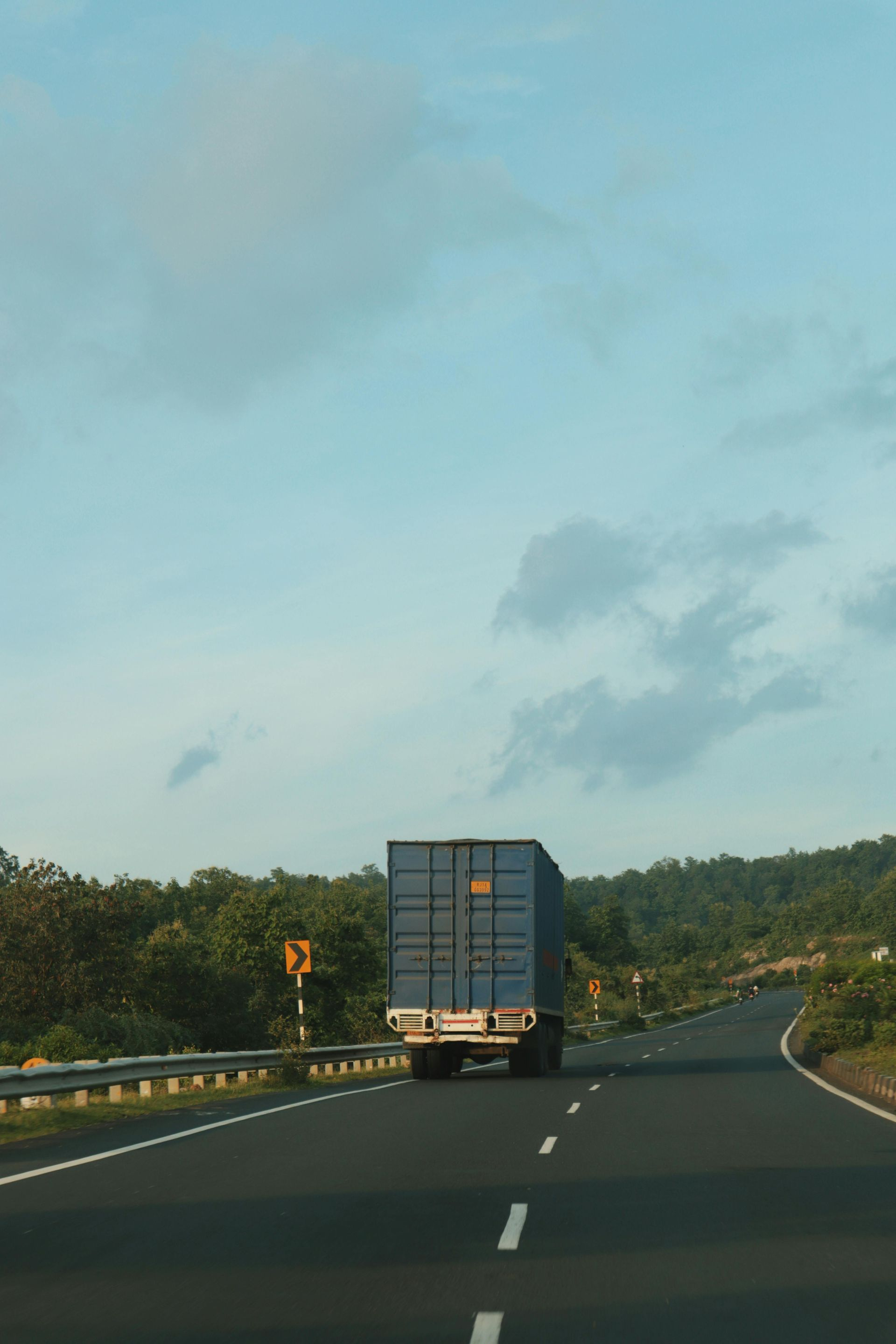 A semi-truck with a blue container travels on a highway. Green trees line the roadside, and the sky is blue.