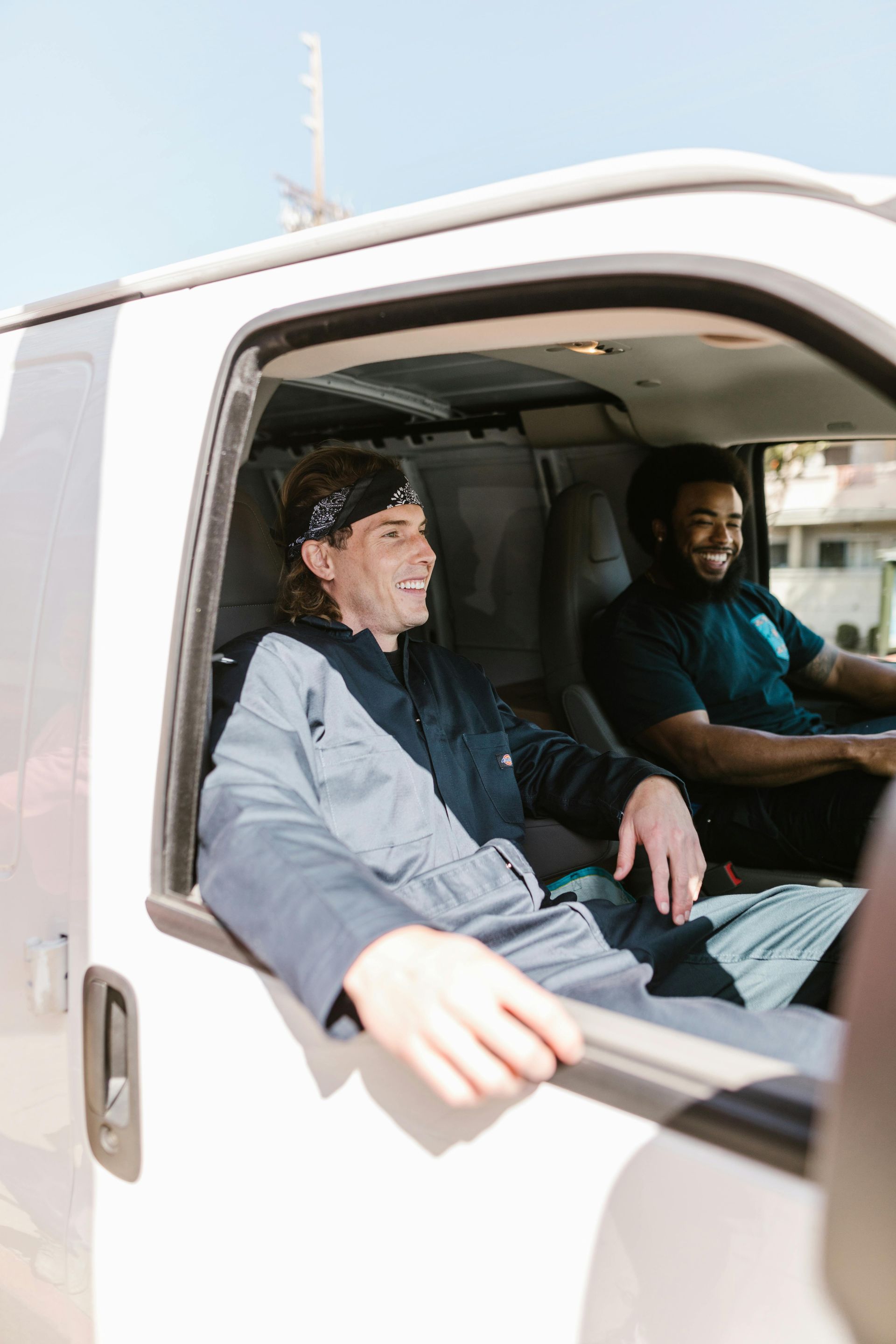 Two men in a white van, smiling. One wearing a bandana and overalls, the other a dark shirt. Sunlight.