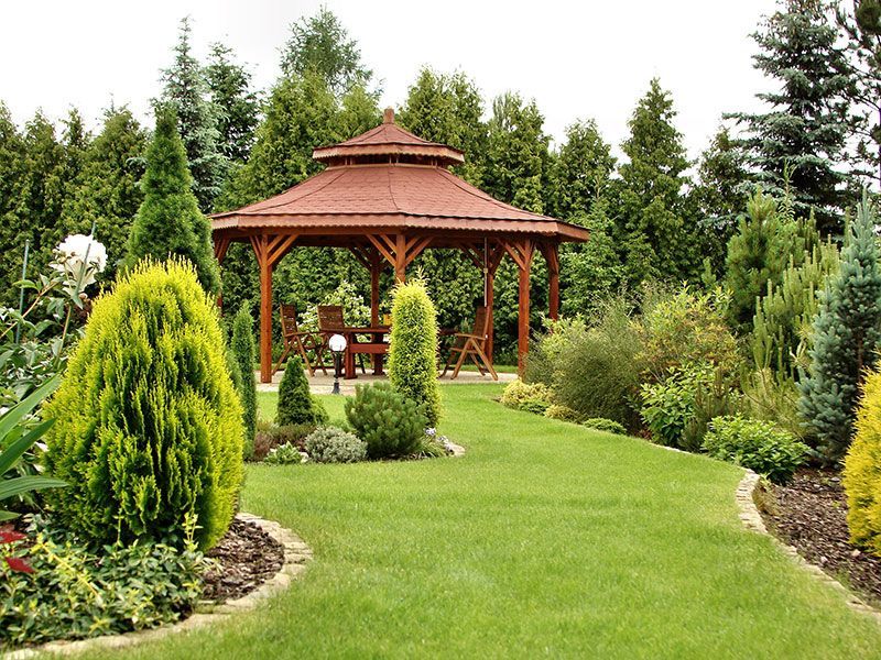 A gazebo with a brown roof in a lush green garden with various trees.