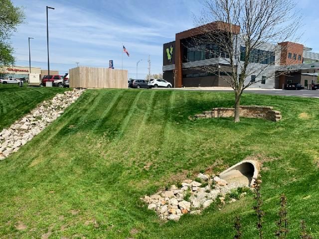 Grassy slope with drainage pipe, rocks, and a modern building under a blue sky.