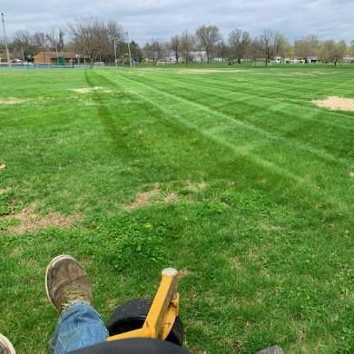 Person mowing a grassy field on a riding lawnmower with stripes. Cloudy sky, buildings in the distance.