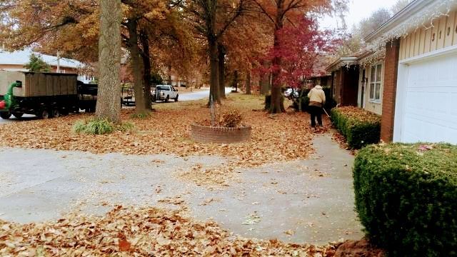 Leaves on the ground; people raking in front of a house. Trees with fall colors, a truck.