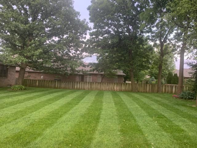 Lawn with alternating green stripes, trees in background, wooden fence, brick building.