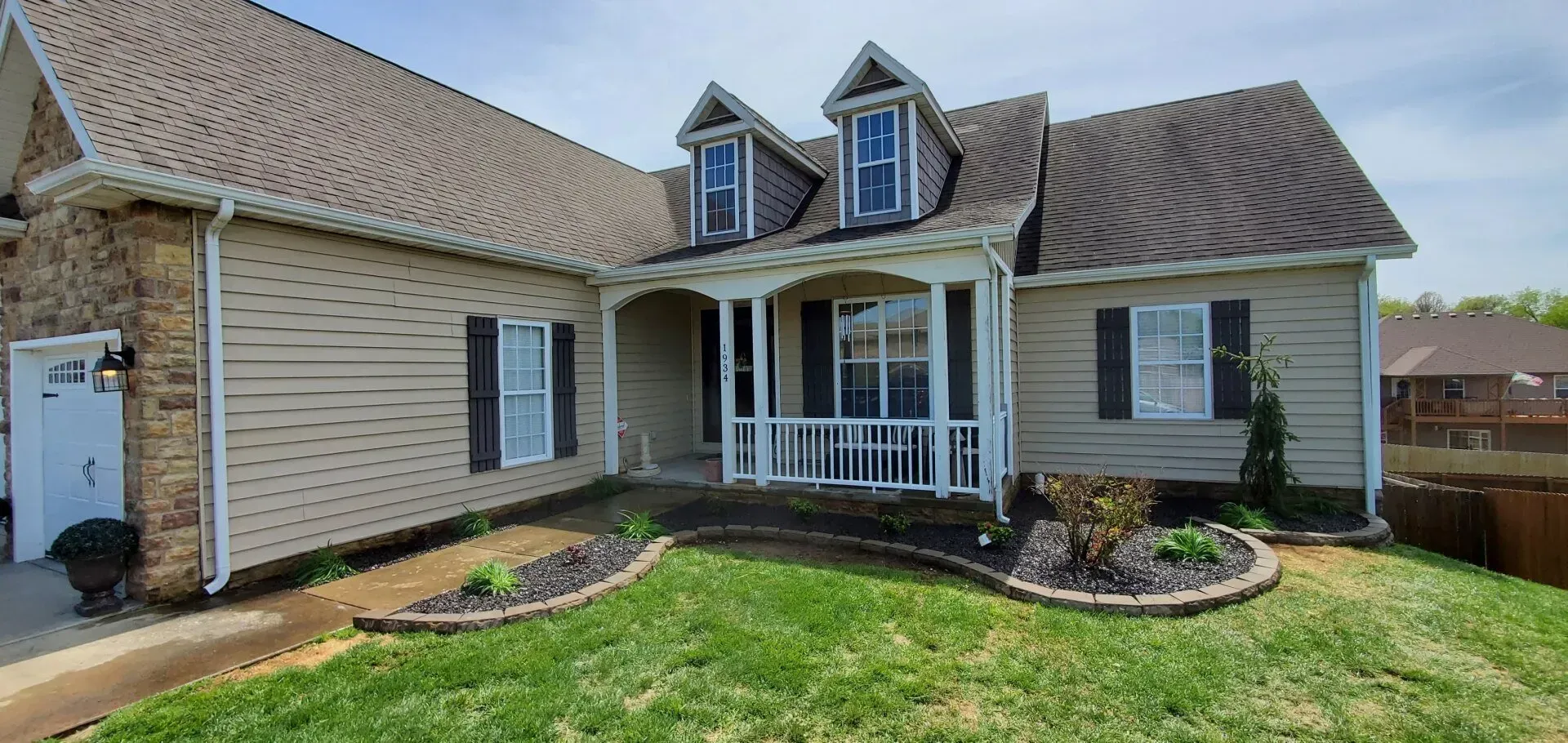 Tan house with brown roof, black shutters, and white porch. Green grass with flower beds.