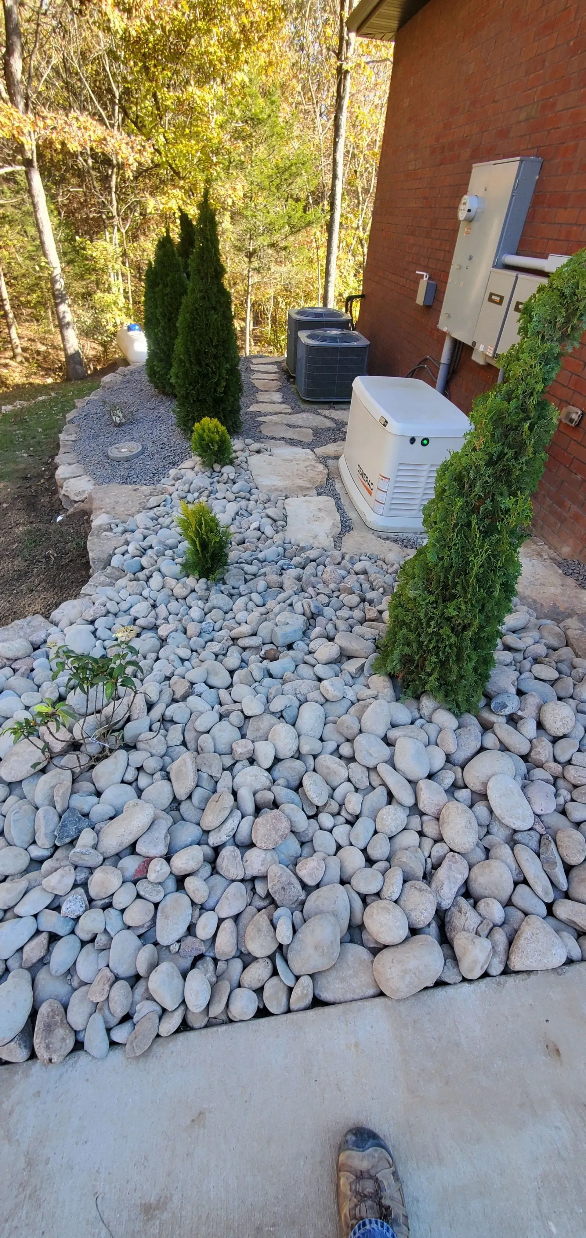 A rock garden with small green shrubs and trees alongside a building with outdoor appliances.