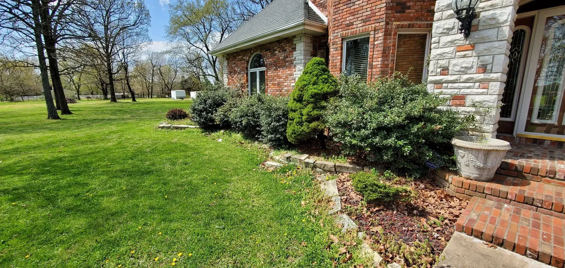 A brick house with a green lawn and bushes in front. A flower pot sits on the porch.
