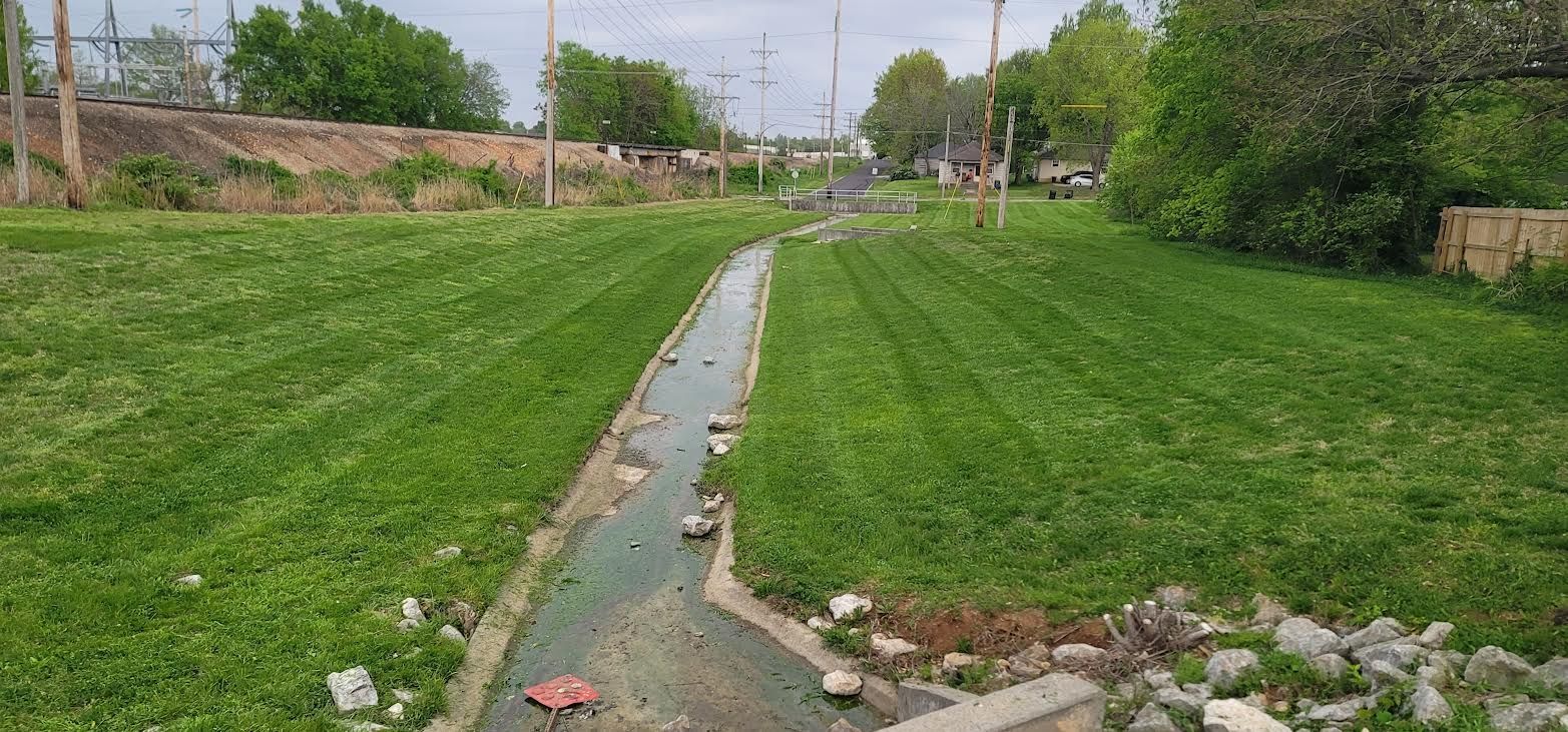 A grassy ditch with a stream of water in the middle. Trees and houses are in the background.