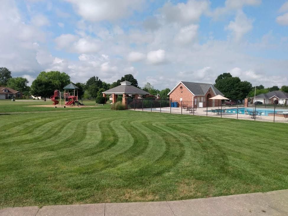 Green lawn with wave-like mowing pattern. Structures and a pool are in the background, under a cloudy sky.