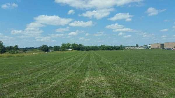 Green field of mown grass under a blue sky with fluffy white clouds; trees and a building are in the distance.