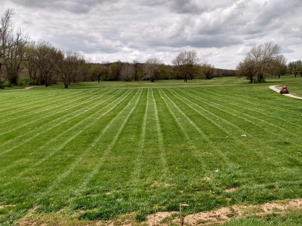 Lawn mowed in straight stripes across a grassy field under a cloudy sky. Trees and a small vehicle are in the background.