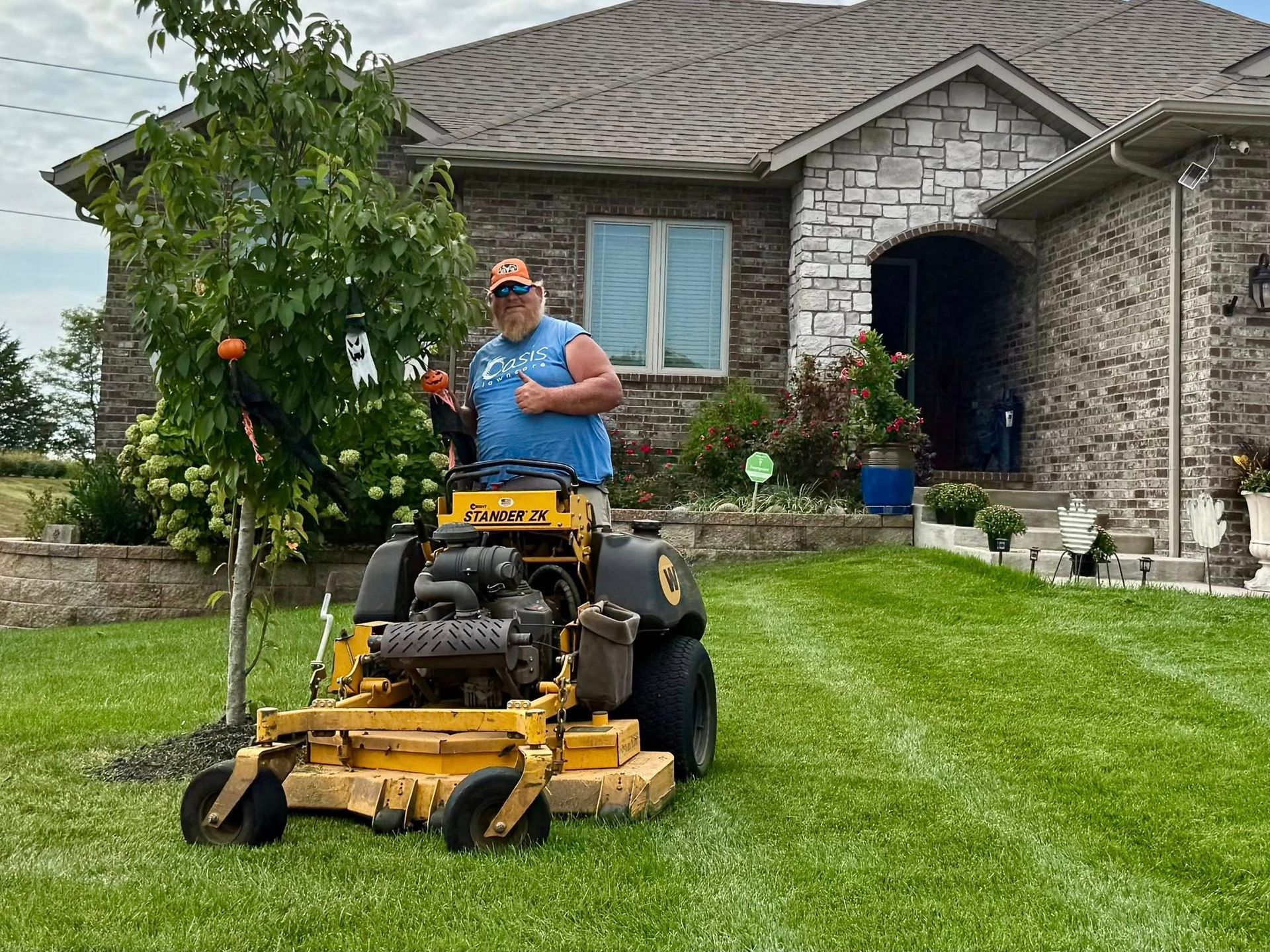 Man on yellow zero-turn mower cutting grass in front of a brick house with a stone front.